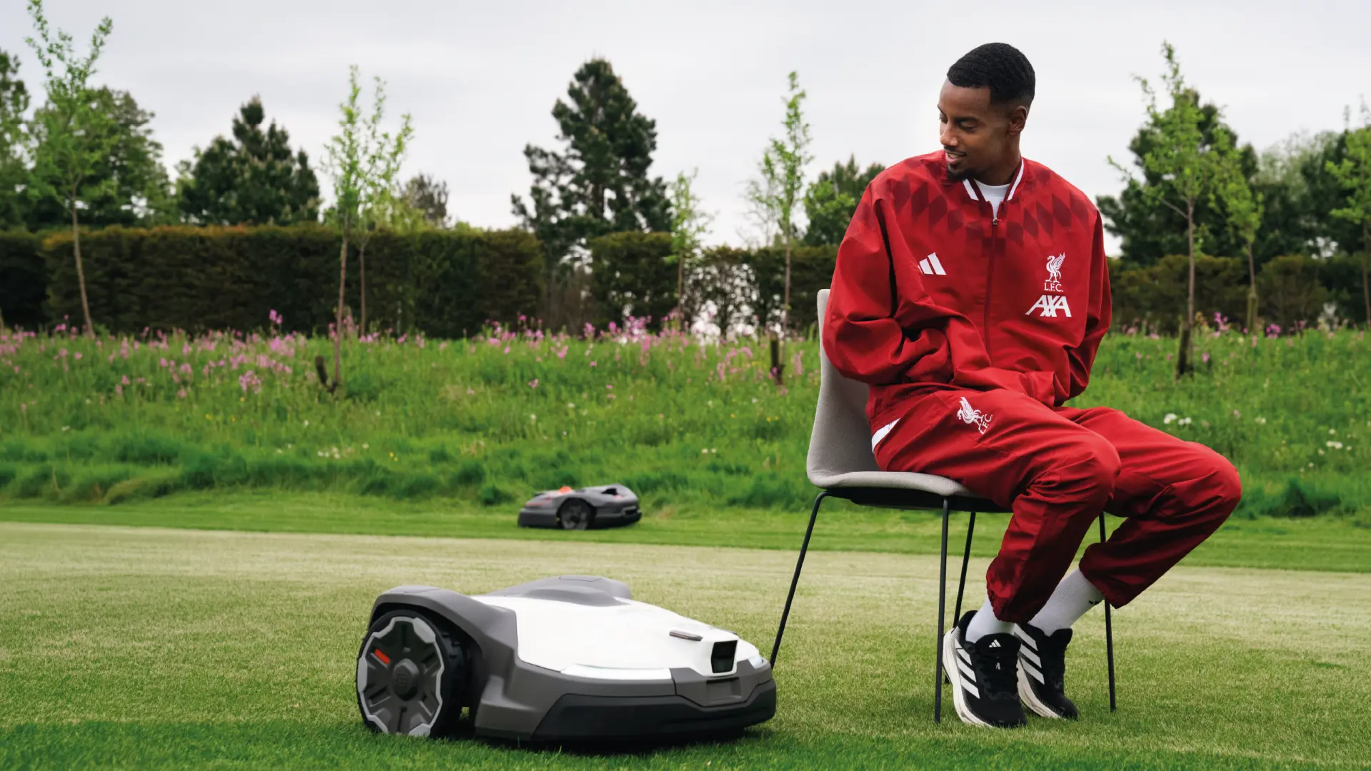 Alexander Isak in a Liverpool FC tracksuit sitting on a chair beside Husqvarna robotic lawnmowers